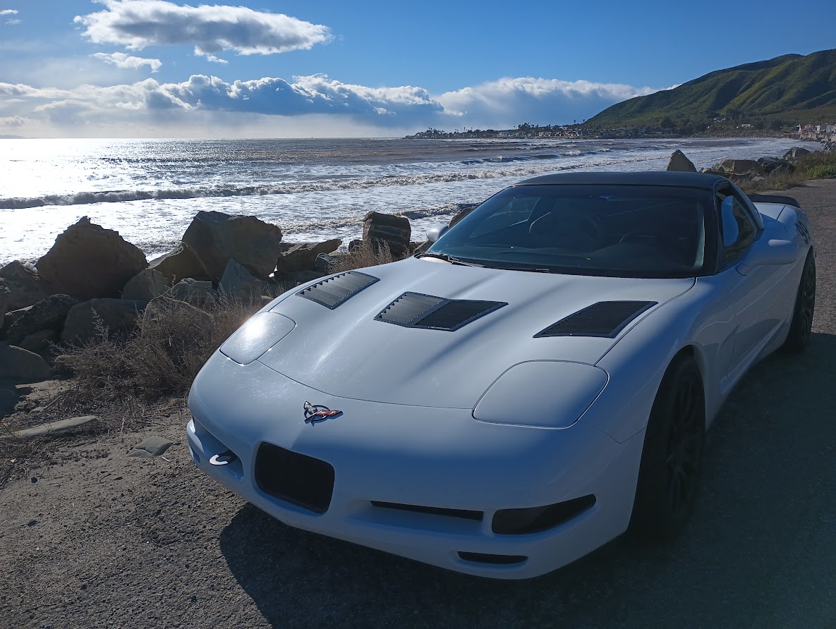 A white c5 Corvette with black hood vents parked by the beach