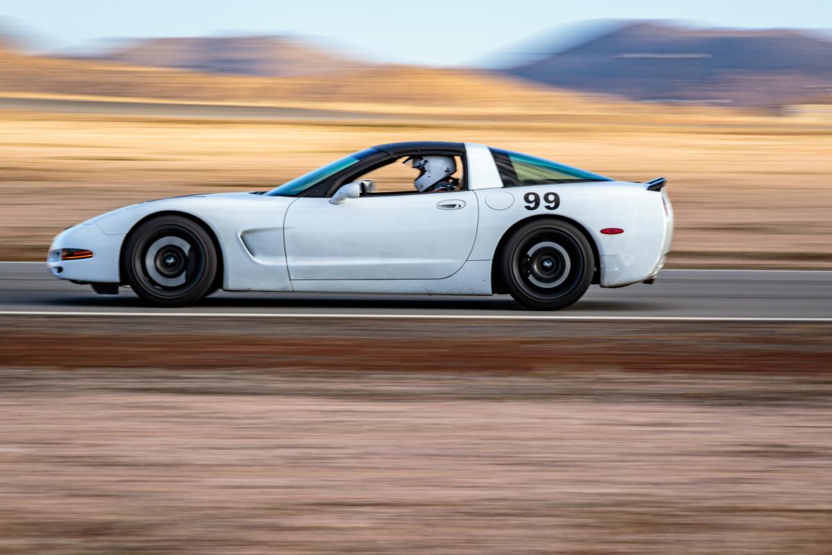 White #99 C5 Corvette driving at the track, background is blurry showing rapid motion, and the car is sitting lower than stock on coilovers